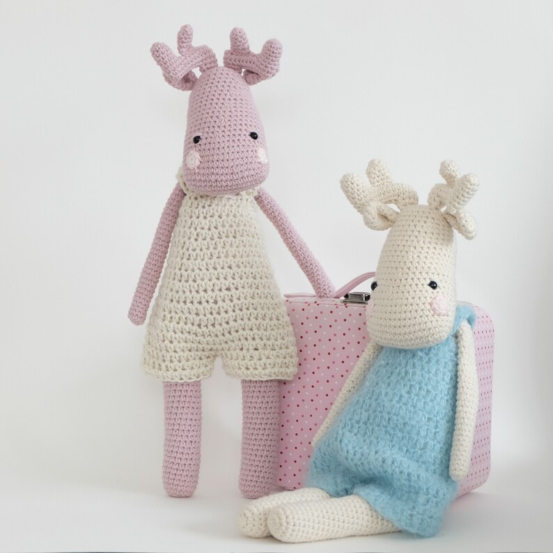 May include: Two crocheted reindeer toys, one pink and one white with blue clothing, sitting next to a pink and white polka dot suitcase.