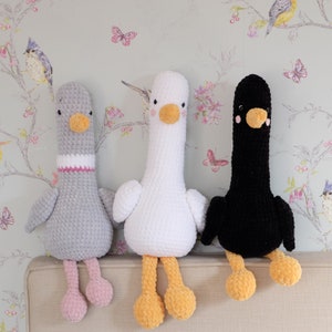 May include: Three crocheted stuffed animals shaped like birds. The bird on the left is gray with pink feet, the middle bird is white with yellow feet, and the bird on the right is black with yellow feet.