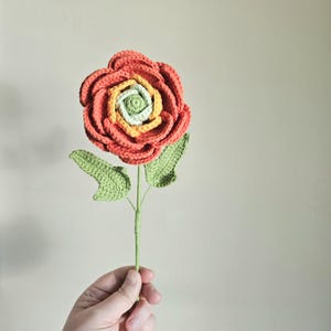 Könnte beinhalten: Handgefertigte Häkelblume in leuchtenden Farben. Die Blume hat Schichten aus korallenfarbenem, gelbem und hellgrünem Garn, mit grünen Blättern und einem grünen Stiel. Ein einzigartiges dekoratives Element.