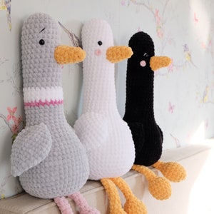 May include: Three crocheted goose plushies, one gray, one white, and one black, sitting on a couch. Each goose has a yellow beak and feet.