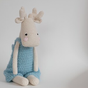 May include: A crocheted stuffed animal moose wearing a blue knitted outfit. The moose has black eyes and pink cheeks.