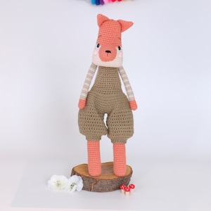 May include: A crocheted fox plush toy wearing brown overalls. The fox has a pink nose and white stripes on its arms. The toy is standing on a wooden log.