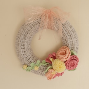Puede incluir: Una corona de ganchillo gris hecha a mano, adornada con flores de ganchillo en tonos rosa, amarillo y melocot&oacute;n. Un delicado lazo de encaje rosa se encuentra en la parte superior. La corona es una pieza decorativa encantadora.