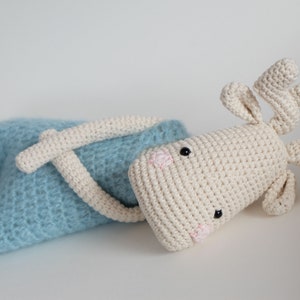 May include: A crocheted stuffed animal in the shape of a reindeer. The reindeer is lying on its side with its head turned to the side. It is wearing a light blue crocheted outfit. The reindeer has black eyes, a pink nose, and brown antlers.
