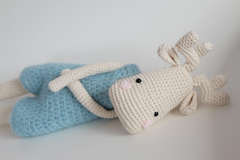 May include: A crocheted stuffed animal in the shape of a reindeer. The reindeer is lying on its side with its head turned to the side. It is wearing a light blue crocheted outfit. The reindeer has black eyes, a pink nose, and brown antlers.