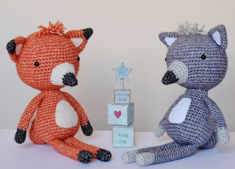 May include: Two crocheted stuffed animals, a fox and a wolf, sitting on a white surface. The fox is orange with a white belly and black nose. The wolf is gray with a white belly and black nose. A small wooden block tower with the words "shining star", "hug me" and a heart is behind the animals.
