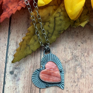 May include: A silver chain necklace with a heart-shaped pendant. The pendant is made of metal with a pink and orange stone in the center.
