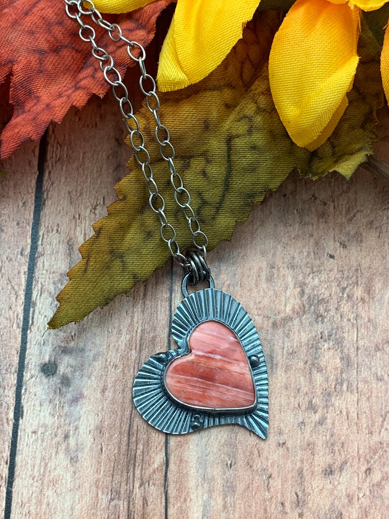 May include: A silver chain necklace with a heart-shaped pendant. The pendant is made of metal with a pink and orange stone in the center.