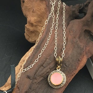 May include: A silver chain necklace with a round pendant. The pendant has a pink center surrounded by a silver border with small, clear stones. The necklace is displayed on a piece of brown wood.