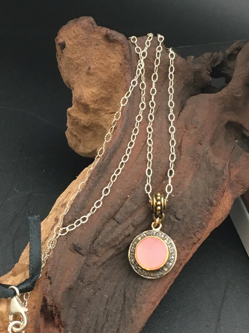 May include: A silver chain necklace with a round pendant. The pendant has a pink center surrounded by a silver border with small, clear stones. The necklace is displayed on a piece of brown wood.
