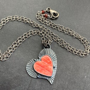 May include: A silver chain necklace with a heart-shaped pendant. The pendant is made of silver and has a pink stone in the center. The stone has a striated pattern.