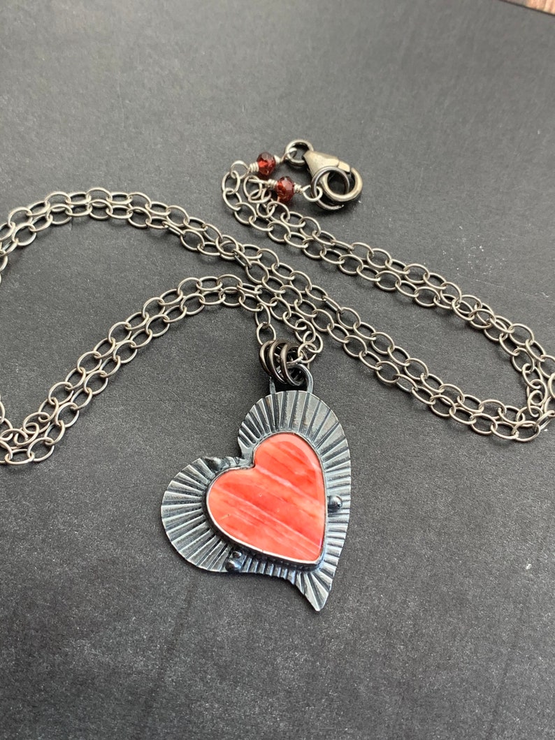 May include: A silver chain necklace with a heart-shaped pendant. The pendant is made of silver and has a pink stone in the center. The stone has a striated pattern.