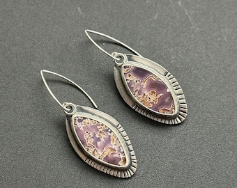 May include: A pair of silver earrings with a leaf-shaped design. Each earring features a purple and tan stone set in a silver bezel with a textured border. The earrings have a hook closure.