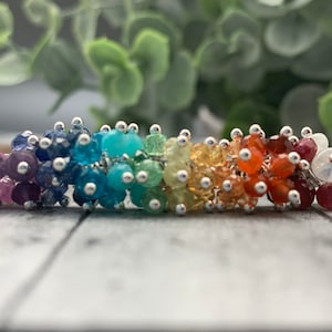 May include: A rainbow-colored gemstone bracelet with silver wire and beads. The bracelet features a variety of gemstones, including amethyst, sapphire, turquoise, peridot, citrine, carnelian, ruby, and moonstone.