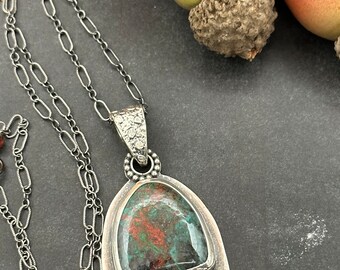 May include: A silver pendant necklace with a green and red gemstone, surrounded by a silver frame. The pendant is accented with a cluster of small red gemstones.