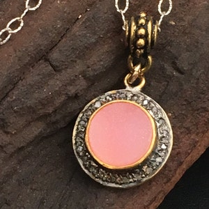 May include: A silver chain necklace with a round pendant. The pendant has a pink center, a gold border, and a ring of small, dark stones. The necklace is displayed on a dark, textured surface.