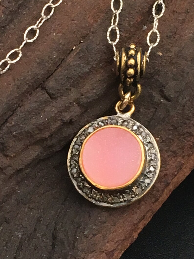 May include: A silver chain necklace with a round pendant. The pendant has a pink center, a gold border, and a ring of small, dark stones. The necklace is displayed on a dark, textured surface.