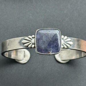 May include: A silver cuff bracelet featuring a large, square, dark blue gemstone at the center. The bracelet has a floral design on each side of the gemstone. The cuff is open at the bottom.