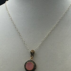 May include: A silver chain necklace with a round pendant. The pendant features a pink center stone surrounded by a dark metal frame with small, sparkling accents. The necklace is displayed on a white mannequin.
