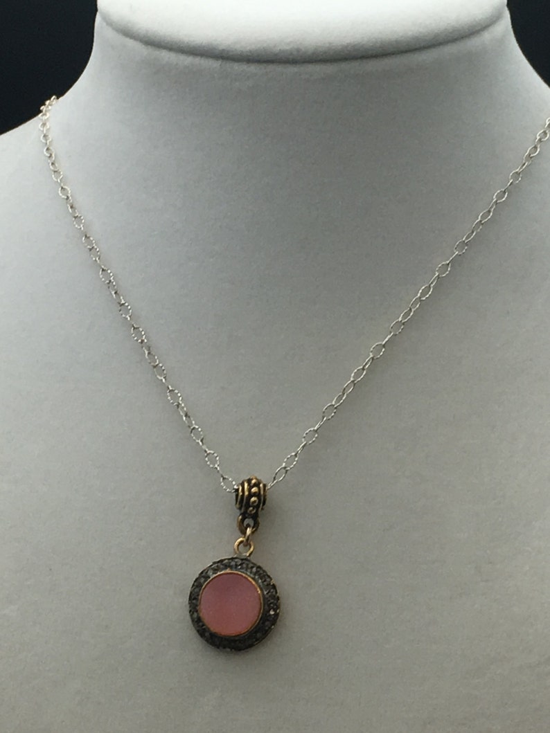 May include: A silver chain necklace with a round pendant. The pendant features a pink center stone surrounded by a dark metal frame with small, sparkling accents. The necklace is displayed on a white mannequin.