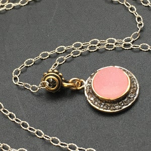 May include: A silver chain necklace with a round pendant. The pendant has a pink center stone, a gold-colored bezel, and a border of small, dark stones. The necklace is displayed on a dark surface.