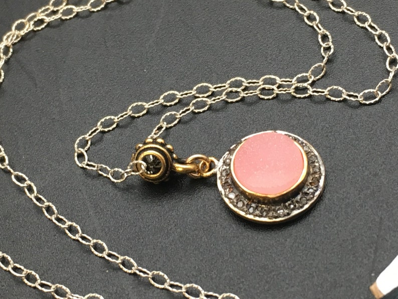 May include: A silver chain necklace with a round pendant. The pendant has a pink center stone, a gold-colored bezel, and a border of small, dark stones. The necklace is displayed on a dark surface.