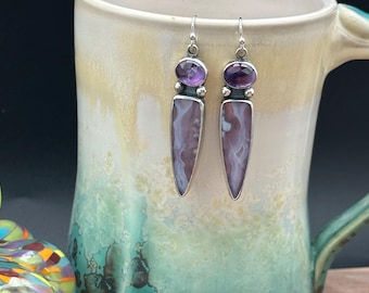 May include: A pair of silver earrings with a purple amethyst stone at the top and a long, rectangular, purple and white stone below. The earrings are displayed in a ceramic mug with a turquoise and cream glaze.