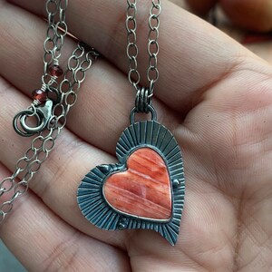 May include: A silver chain necklace with a heart-shaped pendant. The pendant features a red and orange stone with a silver border.
