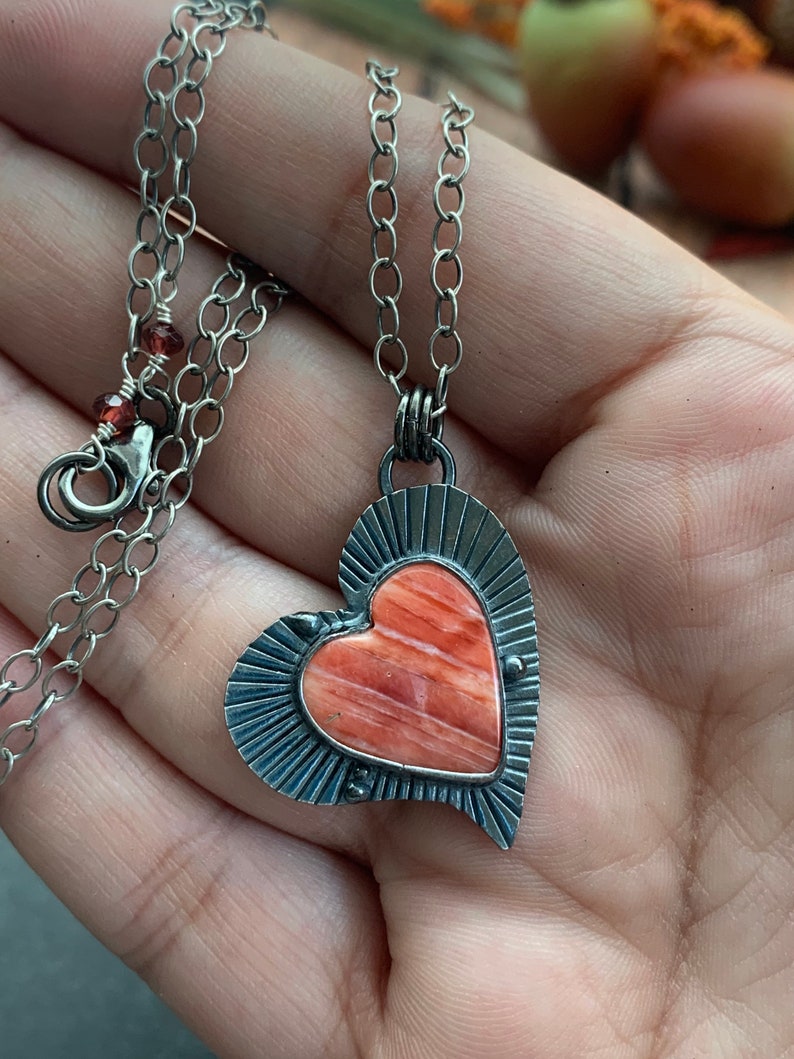 May include: A silver chain necklace with a heart-shaped pendant. The pendant features a red and orange stone with a silver border.