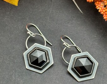 May include: A pair of silver-toned earrings. Each earring features a black, faceted, hexagonal gemstone set within a hexagonal frame. The earrings have a simple, elegant design with a hook closure.