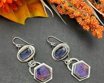 May include: A pair of silver earrings with purple and blue gemstone accents. The earrings feature a hexagonal shaped stone with a silver frame and a smaller oval shaped stone with a silver frame.