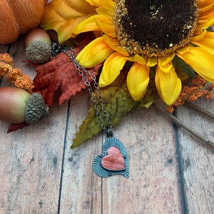 May include: A silver heart-shaped pendant necklace with a pink and orange stone inlay. The necklace is displayed on a wooden surface with fall-themed decorations.