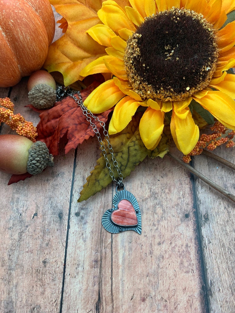May include: A silver heart-shaped pendant necklace with a pink and orange stone inlay. The necklace is displayed on a wooden surface with fall-themed decorations.
