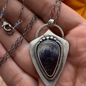May include: A silver pendant necklace with a dark blue gemstone set in a silver shield-shaped frame. The pendant is attached to a silver chain with a clasp.