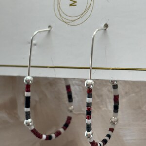 May include: A pair of silver-toned hoop earrings featuring a repeating pattern of small red, white, black, and grey beads, accented with larger smooth silver-toned spherical beads. The earrings are attached to a white display card with a gold letter M.