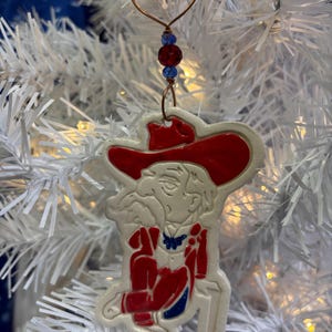 May include: A white ceramic Christmas ornament depicting a cartoon figure wearing a red hat and red and blue clothing. The ornament is hanging on a white artificial Christmas tree.