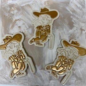 May include: Three identical ceramic ornaments featuring a stylized figure wearing a wide-brimmed hat and holding a cane. The design is finished in a metallic gold paint against a cream-colored background, with a small hole at the top for hanging.