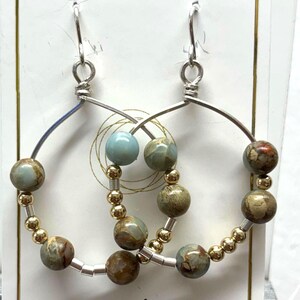 May include: Silver hoop earrings with earth-toned beads and gold accents. The earrings feature a mix of round, patterned beads and smaller gold beads. The card behind the earrings reads "LOVE" and "Melony Designs."