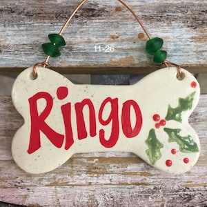 May include: White ceramic bone-shaped ornament with the name "Ringo" written in red paint. The ornament has green holly leaves and red berries painted on it. It has a copper wire hanger with green glass beads.