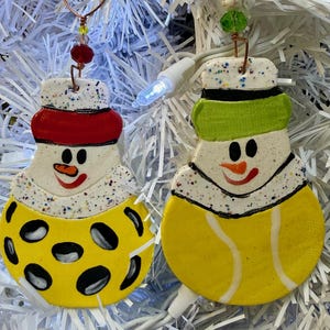 May include: Two snowman ornaments with speckled white heads, orange carrot noses, and black eyes. One snowman wears a red hat and a yellow body with black spots. The other wears a green hat and a yellow body with white tennis ball lines.