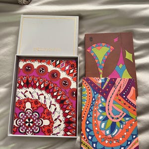 May include: A set of three rectangular notebooks with vibrant, multi-colored geometric and floral patterns. One notebook is inside a white box labeled EMILIO PUCCI, while two others are placed beside it, featuring intricate designs and signatures.
