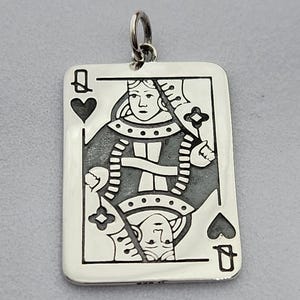 May include: Silver pendant in the shape of a playing card, featuring the Queen of Hearts. The card design includes the suit symbols and the letter "Q". The pendant has a loop at the top for a chain.