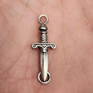 May include: Silver-toned sword charm with a detailed design. The charm has a loop at the top for attaching to a necklace or bracelet.