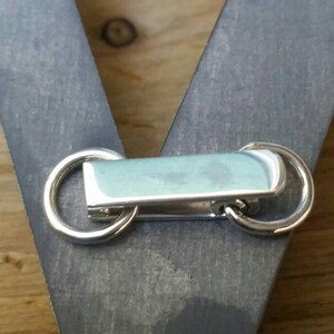 Peut inclure: Gros plan sur un fermoir en m&eacute;tal argent&eacute; avec deux anneaux circulaires et une pi&egrave;ce centrale rectangulaire bleu clair. Le fermoir est fix&eacute; &agrave; une surface en bois grise, avec un fond en bois.