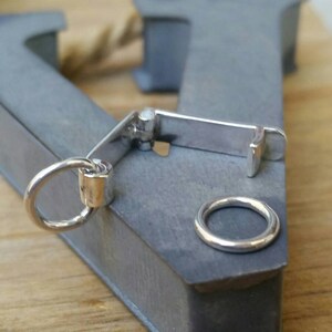 Peut inclure: Un fermoir en m&eacute;tal argent&eacute; avec un petit anneau attach&eacute;, &agrave; c&ocirc;t&eacute; d'une simple bague en argent. Le fermoir est mont&eacute; sur une surface m&eacute;tallique grise, avec une surface en bois en arri&egrave;re-plan.