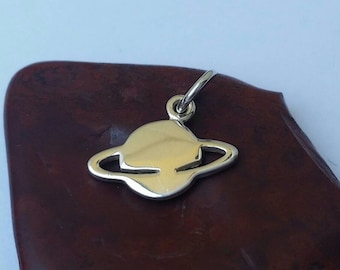 May include: A silver-colored planet charm with a ring around it, suspended from a small loop. The charm is set against a dark brown surface, with a light background.