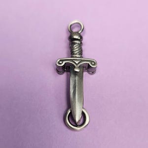 May include: A silver-toned dagger charm with a detailed design. The charm has a loop at the top for attaching to a necklace or bracelet.