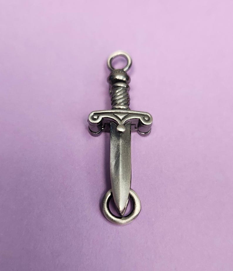 May include: A silver-toned dagger charm with a detailed design. The charm has a loop at the top for attaching to a necklace or bracelet.