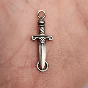 May include: Silver-toned sword charm with a detailed design. The charm has a loop at the top for attaching to a necklace or bracelet.