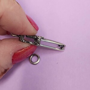 May include: Silver-toned metal clasp with a decorative design. The clasp is open and a small silver-toned metal ring is lying next to it.
